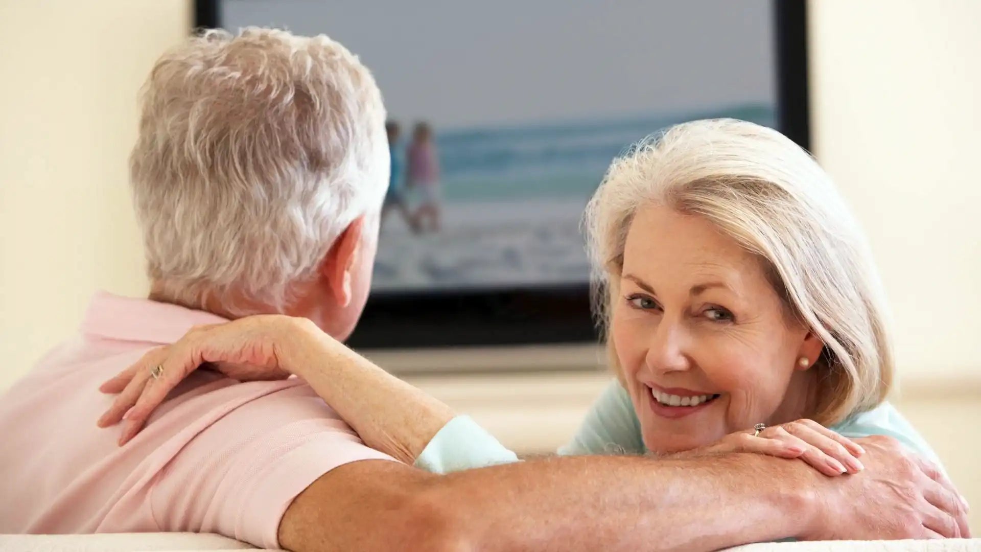 Une couple de personnes seniors regarde la télé dans un canapé et la femme se retourne et sourit.